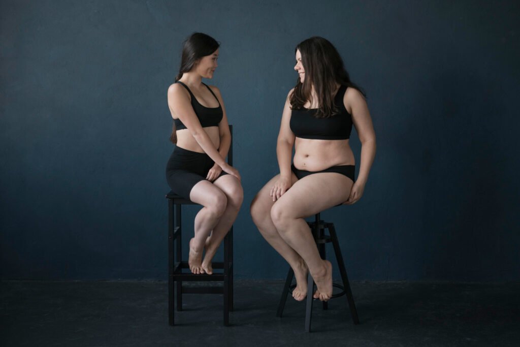Duas mulheres de biotipos diferentes sentadas, ilustrando a comparação de corpos e a possível presença de Lipedema em pernas e braços, mesmo em diferentes pesos.