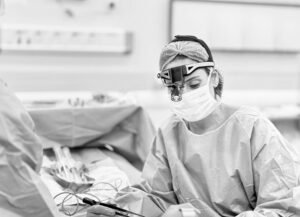 Foto em preto e branco de uma cirurgiã plástica Ana Borba concentrada, usando máscara, gorro e lupas cirúrgicas com foco de luz, durante um procedimento